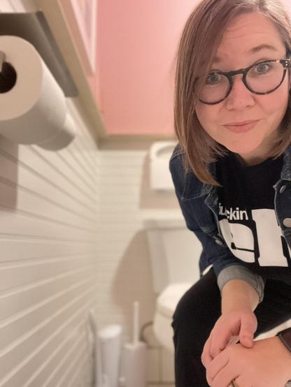 Awkward woman with brown hair and glasses squatting by a toilet in a public restroom.